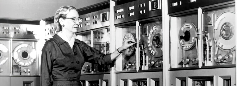 A imagem mostra foto de Grace Hopperusando um vestido longo, de mangas compridas, de cor escura. Ela está segurando uma folha de papel e do lado de uma parede com diversos equipamentos.